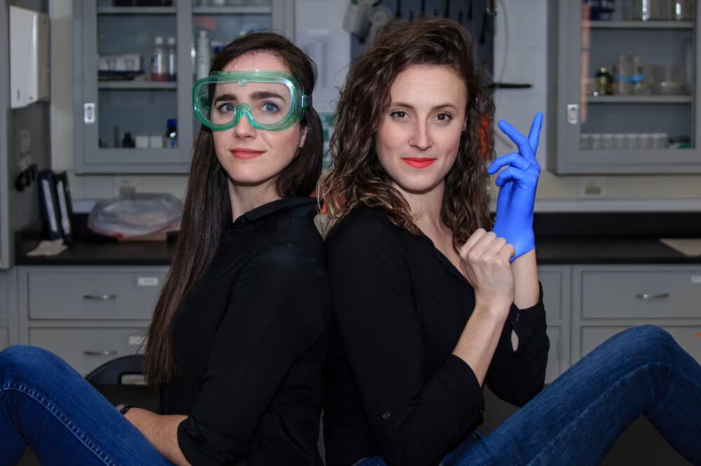 Two women sitting on a lab table. Both are wearing black with jeans. The one on the left is wearing safety goggles, the one on the right is putting on a blue glove.