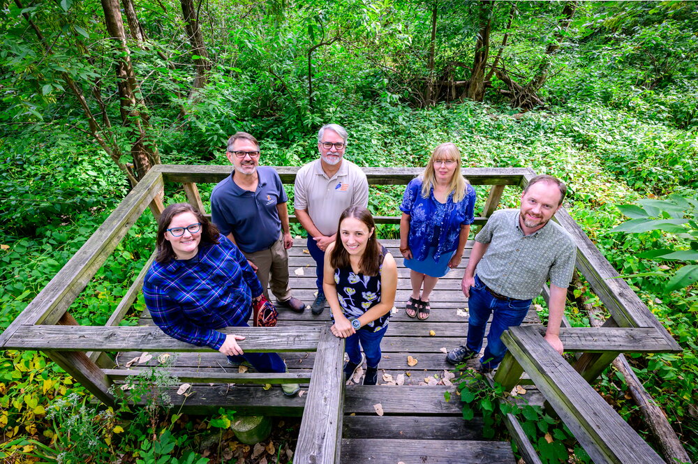 A group of people standing on a wooden platform with woods behind them