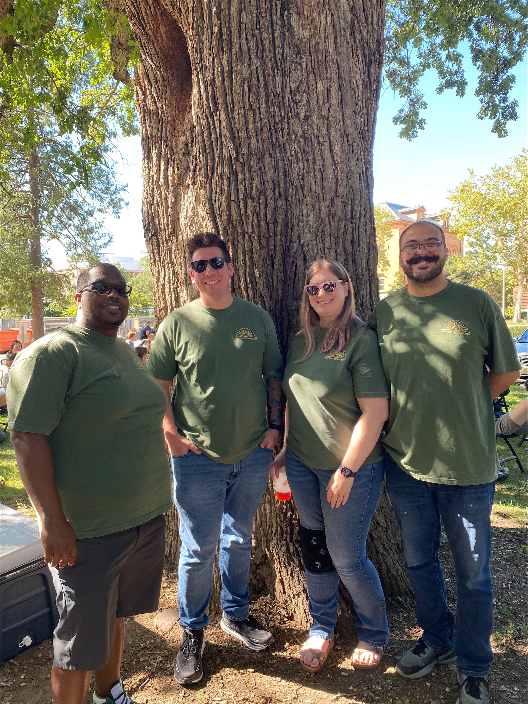 Three men and a woman all in green shirts standing in front of a tree