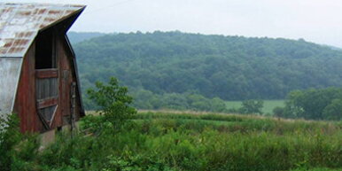 An old barn on the right with a field in the foreground and mountains in the background