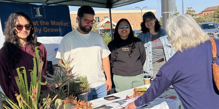 A group of people behind a table smiling at the camera while also talking to a woman at their table