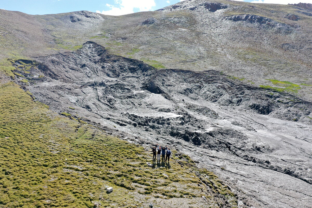 A mix of grassy and rocky hillside with people smiling and waving at the camera 