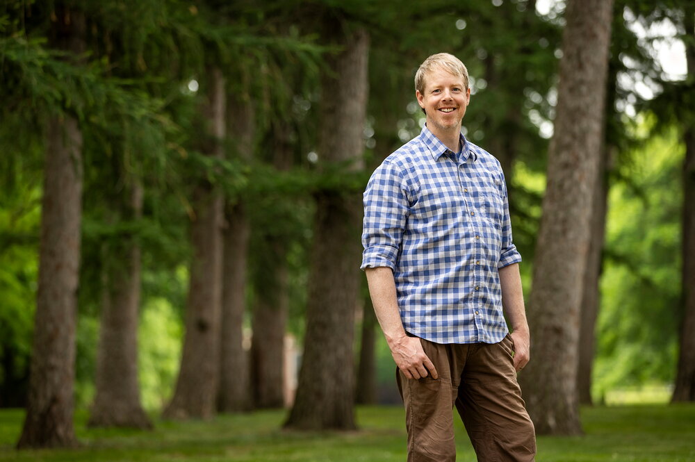 A man in a blue plaid shirt and brown pants standing in front of trees