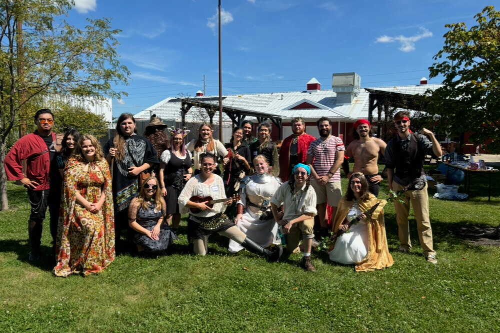 A group of people standing and kneeling in grass wearing fantasy cosplay, with a building behind them