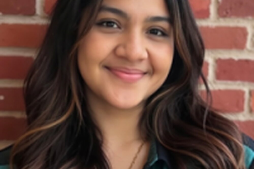 A smiling woman in a green and black flannel standing in front of a brick wall
