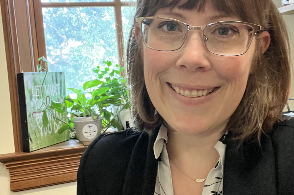 A woman in glasses with a black blazer and white and black flowery shirt smiling in front of a window