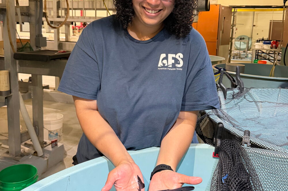 A smiling woman holding a small paddlefish over a large tub with others swimming inside