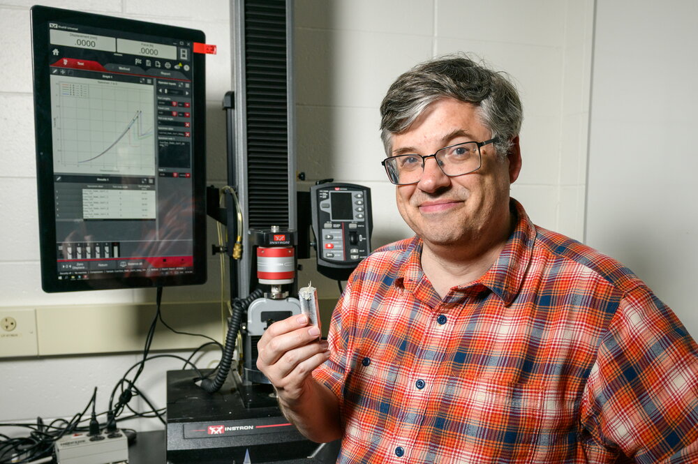 A man smiling in an orange and blue button up in a lab holding an object