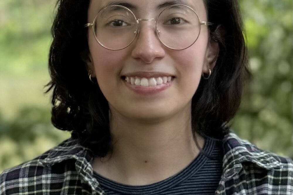 A smiling woman in round glasses and a plaid shirt standing outside