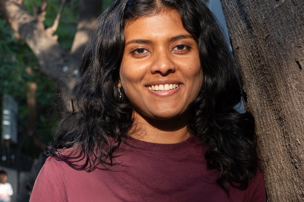 A smiling woman in a maroon shirt standing next to a tree