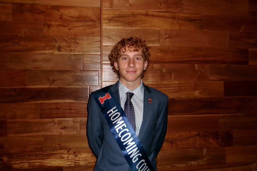 A smiling man in a suit wearing a sash standing in front of a wooden wall
