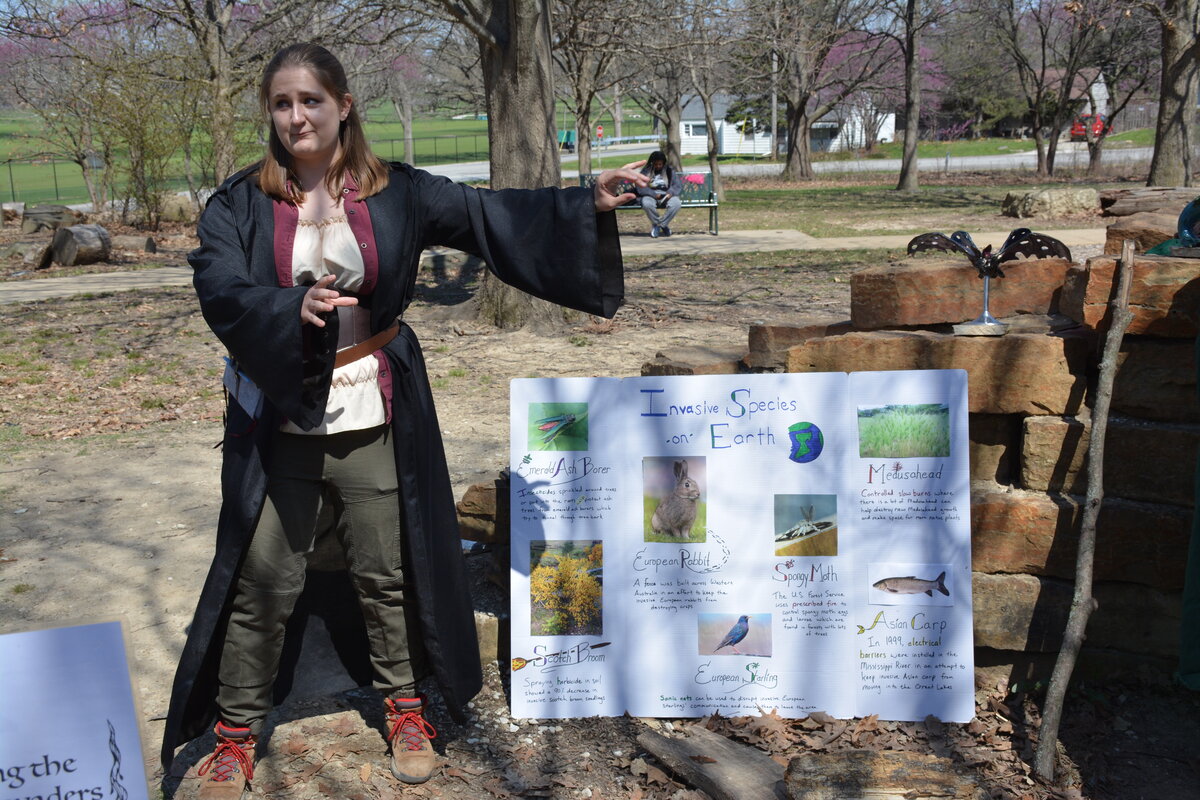 A woman dressed in fantasy garb gesturing to a poster