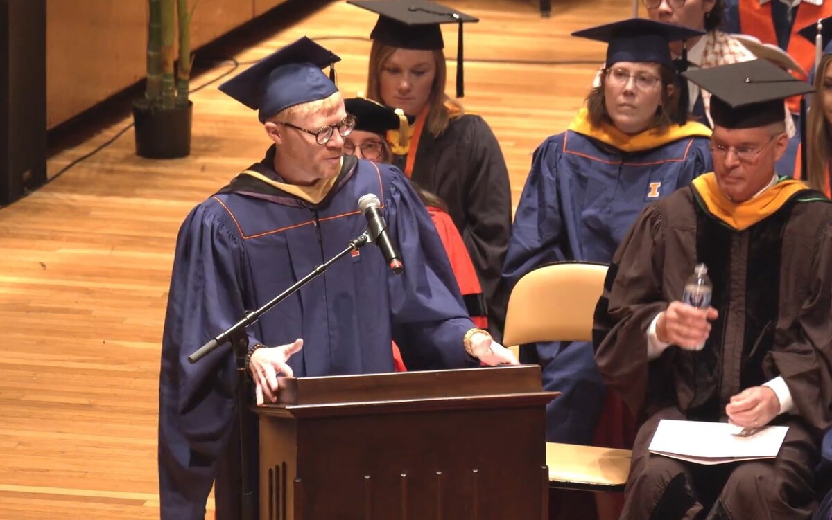 A photo of a man in commencement garb speaking at a podium