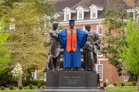 A view of the Alma Mater wearing graduation regalia with the Illini Union in the background. 