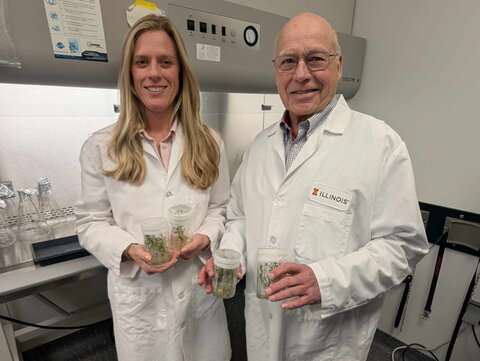  man and a woman in white lab coats hold containers with young green plants in a lab setting.