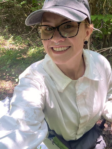 A person in a hat and glasses wearing a white button up shirt taking a selfie outside