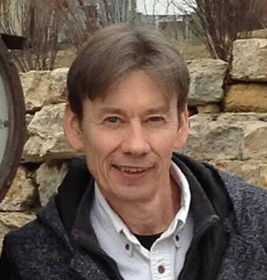 A man in a coat and white button down shirt in front a stone wall