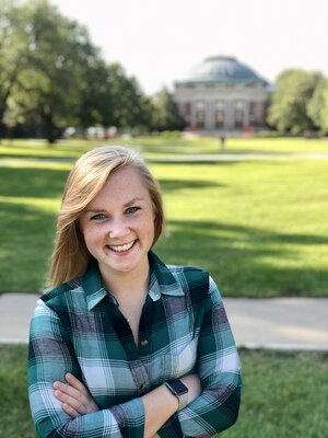 A woman in a blue plaid shirt smiling with a grass and a dome building in the background