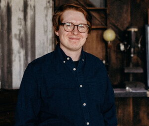 A man in a dark blue button up shirt in glasses with a rustic background