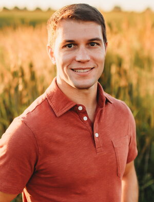 A man in an reddish orange polo smiling with a field behind him