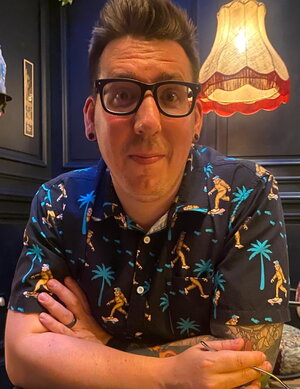 A man in a black polo and black framed glasses with a dark wall and light behind him