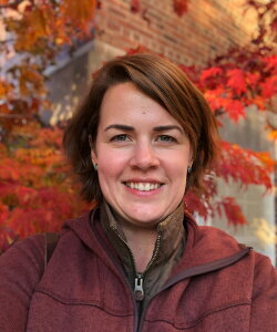 a woman in a maroon jacket standing in front of fall foliage