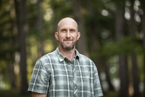 A bald man in a green plaid shirt standing in the woods