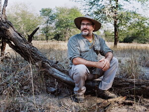 A man in a large hat and safari gear sitting on a downed tree