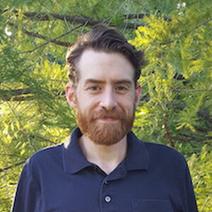 A man with a beard in a blue button down shirt standing in front of green trees
