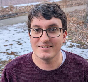 A man with short hair in glasses and a red sweater standing in front of some snowy ground coverage