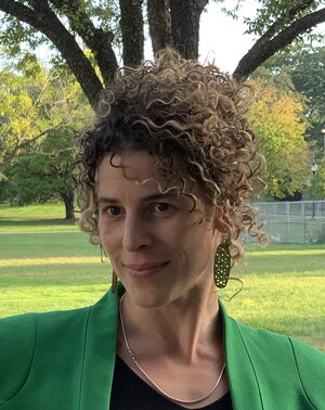 A woman with brown curly hair in a green sports coat with a black top standing in front of a tree and grassy background