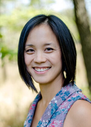 A woman with black hair in a floral shirt and standing in front of a blurry wooded background