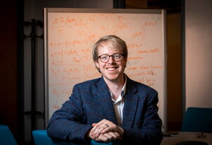 a man with short hair and glasses in a blue blazer and white shirt in front of a white board with orange writing on it