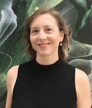 A woman with curly hair in a black sleeveless top standing in front of plants with giant leaves