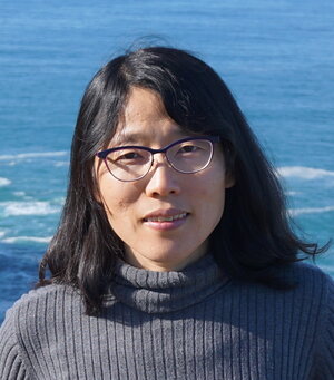 A woman with black hair and glasses in a gray shirt standing in front of the ocean