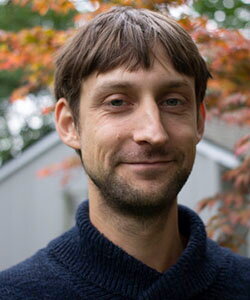 A man with brown hair in a blue button up shirt standing in front of fall foliage on a tree