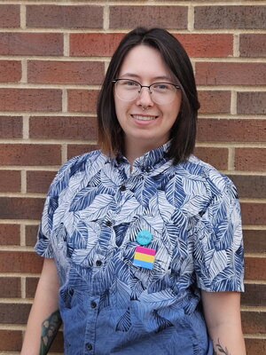A person in glasses with a blue and white plant button up shirt standing in front of a brick wall