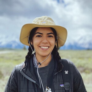 A smiling woman with braided hair in a khaki hat standing outside