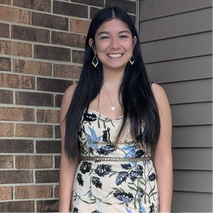 A smiling woman in a dress standing in front of a wall, one half brick and the other siding
