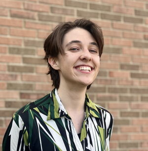 A smiling person in a green and white patterned shirt standing in front of a brick wall