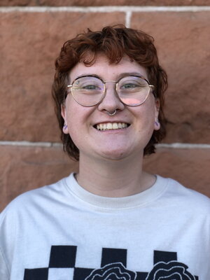 A smiling person with red curly hair wearing glasses and a white t-shirt standing in front of a brick wall