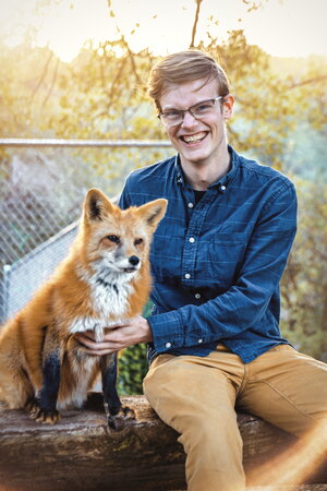A smiling man in a blue button up and khakis sitting outside next to a fox