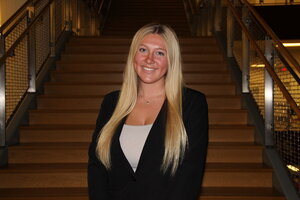 A smiling blonde woman in a suit jacket standing in front of stairs