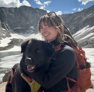 A woman smiling in a snowy valley and hugging a black dog in a harness