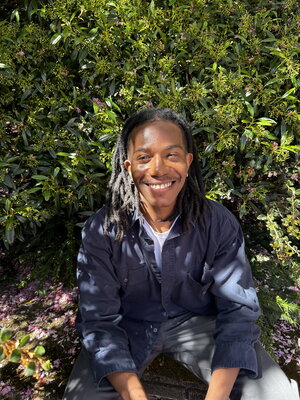 A smiling man in a blue button up shirt sitting in front of some plants