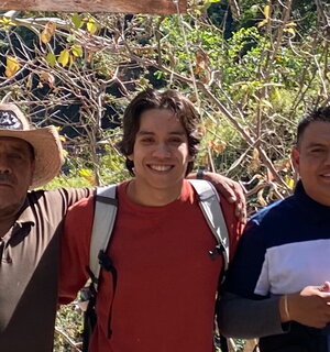 A smiling man in a redshirt and backpack with his arms around two other people outside