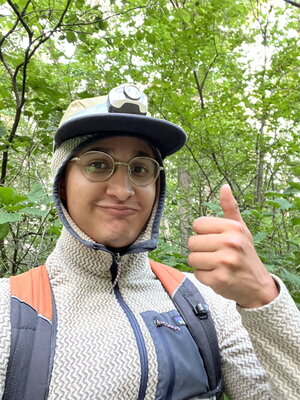 A person in glasses and a jacket giving a thumbs up in the woods
