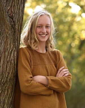 A smiling woman in a brown shirt leaning against a tree