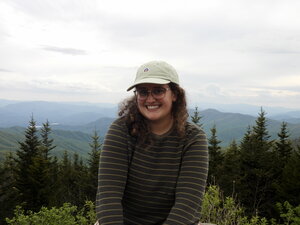 A smiling woman in a hat sitting on a hillside with trees and mountains behind her
