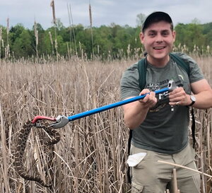 A smiling man in a green shirt, hat, and backpack holding a snake with a grabber